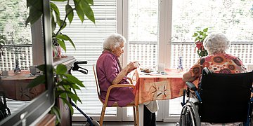 Zwei ältere Frauen sitzen an einem Tisch in einem hellen Raum mit großen Fenstern.