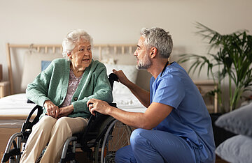 Eine &auml;ltere Dame sitzt im Rollstuhl in ihrem Zimmer. Seitlich von ihr hat sich ein Pfleger hingehockt und unterh&auml;lt sich mit ihr in Augenh&ouml;he. 