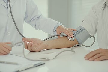 Das Foto zeigt einen Arzt und einen Patienten bei der Blutdruck-Messung vor hellem Hintergrund. Beide sind wei&szlig; gekleidet, K&ouml;pfe und Beine sind nicht zusehen. Beide stehen an einem Tisch. Im Zentrum des Bildes sieht man das Blutdruckmessger&auml;t, dessen am rechten Arm angelegte Manschette und eine Stetoskop in der rechten Armbeuge des Patienten.