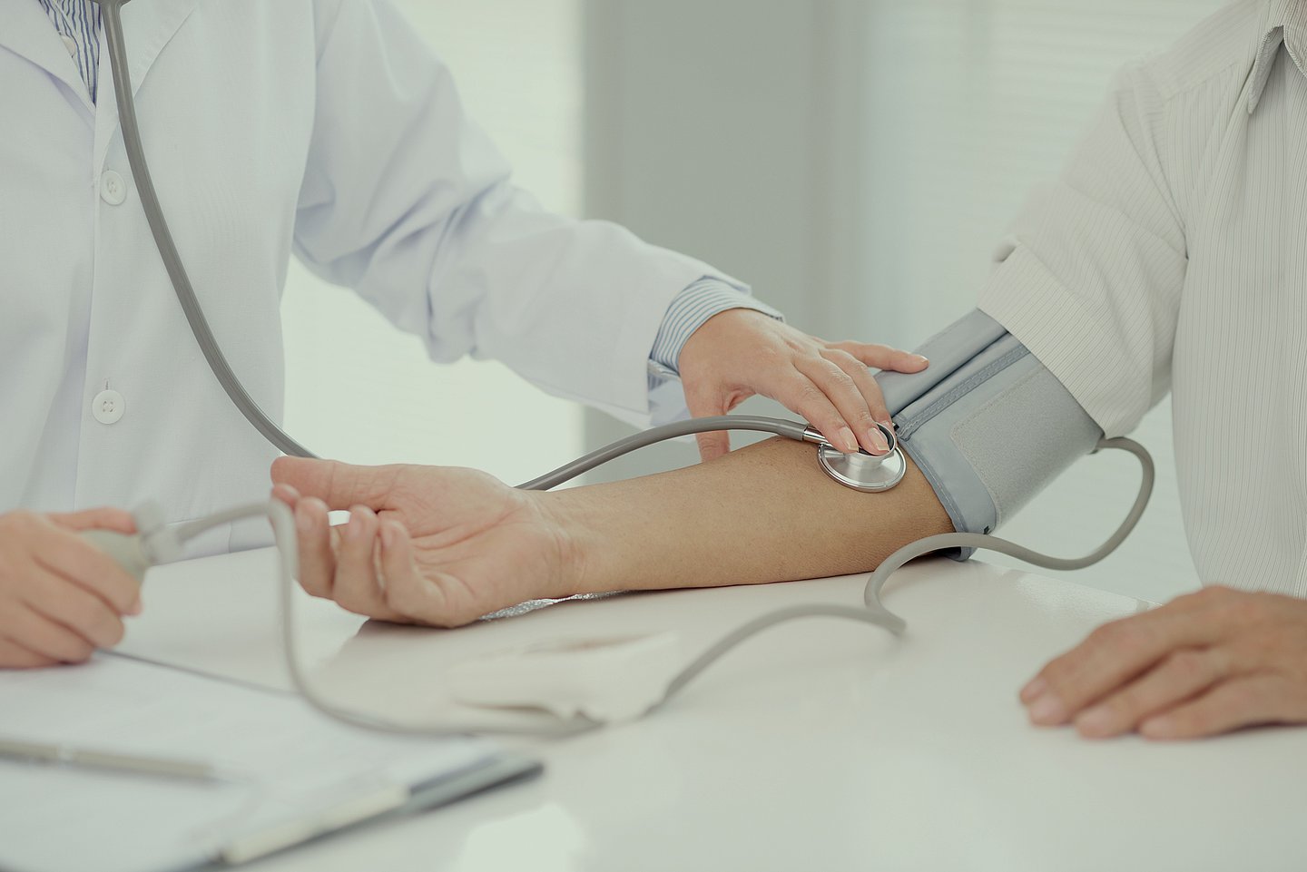Das Foto zeigt einen Arzt und einen Patienten bei der Blutdruck-Messung vor hellem Hintergrund. Beide sind weiß gekleidet, Köpfe und Beine sind nicht zusehen. Beide stehen an einem Tisch. Im Zentrum des Bildes sieht man das Blutdruckmessgerät, dessen am rechten Arm angelegte Manschette und eine Stetoskop in der rechten Armbeuge des Patienten.