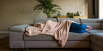Foto: Eine Frau liegt auf einem Sofa und hat ihren Oberk&ouml;rper und ihr Gesicht mit einer Decke verdeckt.