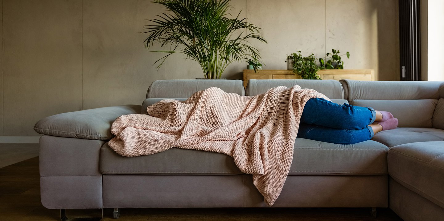 Foto: Eine Frau liegt auf einem Sofa und hat ihren Oberkörper und ihr Gesicht mit einer Decke verdeckt.
