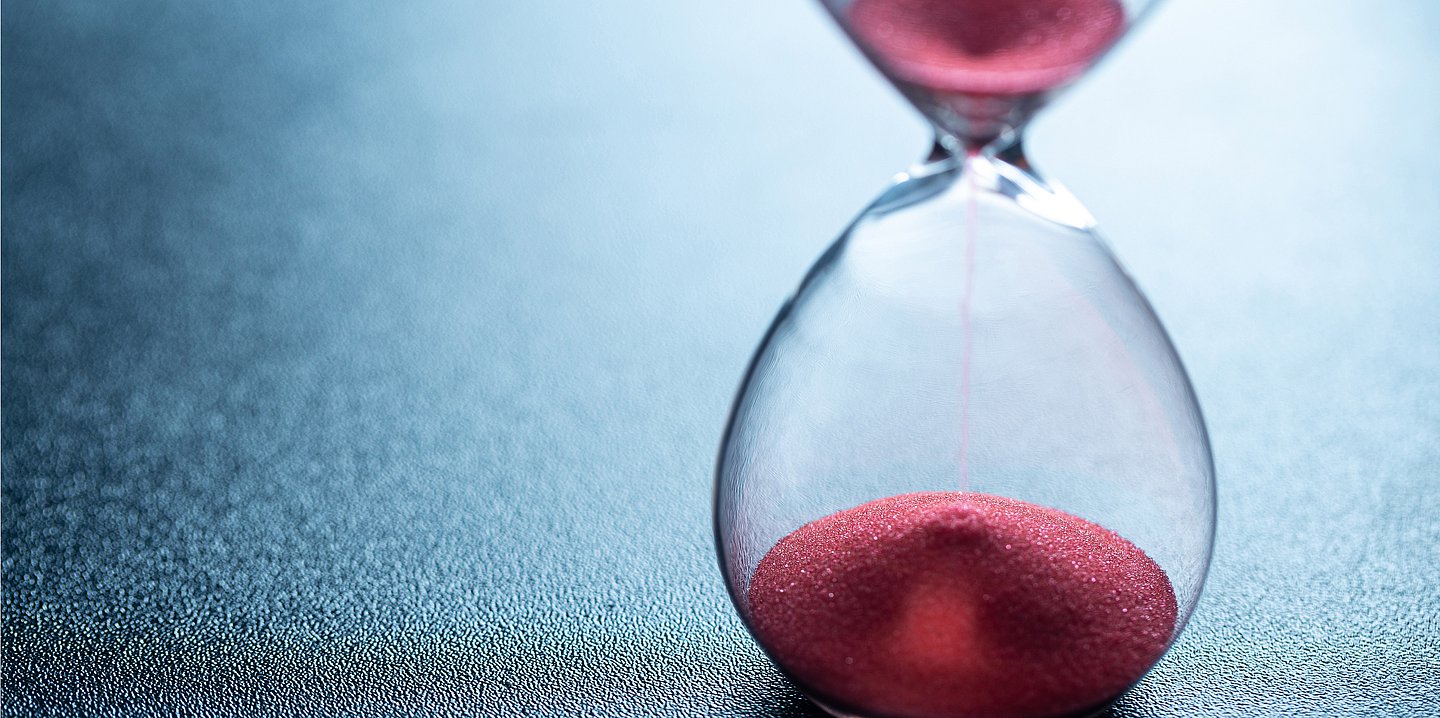 Foto: Roter Sand läuft durch eine Sanduhr.
