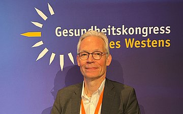 Ein Mann, Matthias Mohrmann, steht vor einer blauen Wand mit dem Schriftzug "Gesundheitskongress des Westens"