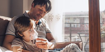 Kind mit Inhalationsmaske Foto: Ein Vater hält seinen Sohn im Arm und hält ihm eine Inhalationsmaske vor das Gesicht.