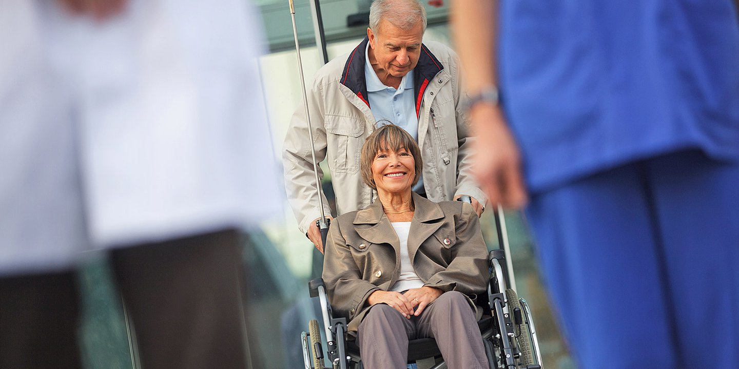 Foto einer fröhlichen älteren Frau, die im Rollstuhl sitzt und von einem älteren Mann aus einem Klinikbereich herausgeschoben wird