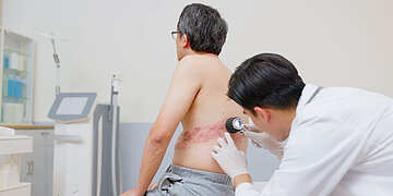Das Bild zeigt einen Patienten mit freiem Oberk&ouml;rper auf einer Liege sitzendend und Ausschlag im seitlich und unteren Bauch- und R&uuml;ckenbereich. Ein Arzt untersucht mit einer beleuchteten Lupe
