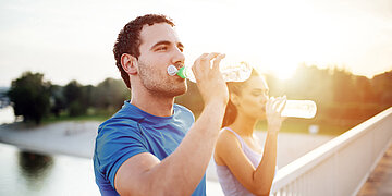 wei sportlich gekleidete Personen trinken im Freien Wasser aus Flaschen; im Vordergrund ein Mann im blauen Shirt, im Hintergrund eine Frau, beide bei warmem Sonnenlicht auf einer Br&uuml;cke oder Promenade.