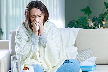 AOK-Gesundheitsbericht erstes Halbjahr 2025 für Westfalen-Lippe Eine Frau sitzt mit einer Decke umhüllt Nase putzend auf der Couch.