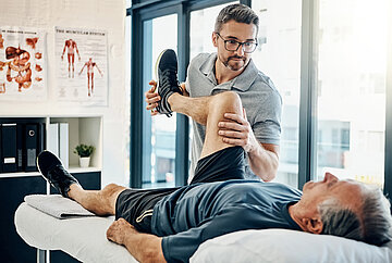 Ein Patient liegt in R&uuml;ckenlage auf der Behandlungsliege beim Physiotherapeuten. Dieser hat Fu&szlig; und Oberschenkel umfasst und das Bein im rechten Winkel vor den Bauch des Mannes positioniert und beobachtet die Reaktionen des Patienten.