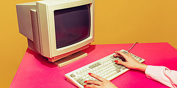 Ein Schreibtisch, auf dem ein alter Monitor steht. Der Tisch ist pink, der Hintergrund ockerfarbig.