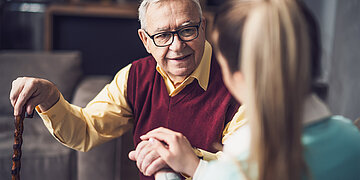Ein &auml;lterer Pflegebed&uuml;rftiger sitzt einer Pflegerin gegen&uuml;ber, die von hinten zu sehen ist.