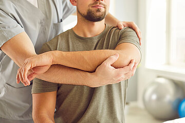 Ein Mann in sitzender Haltung wird von einer hinter ihm stehenden Physiotherapeutin an seiner rechten Schulter und dem Arm behandelt.