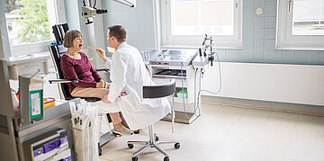 Foto: Patientin sitzt auf einem Behandlungsstuhl, w&auml;hrend ein Arzt ihren Rachen mit einem Holzspatel untersucht.