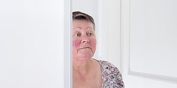 Rosazea Foto: Eine Frau mittleren Alters mit kurzen braunen Haaren und mit geröteten Wangen schaut in einen Spiegel.