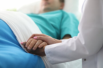 Eine &Auml;rztin in Nahaufnahme h&auml;lt die Hand eines Mannes im Krankenbett.