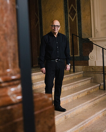 Armin Nassehi steht auf einer breiten Steintreppe in einem historischen Geb&auml;ude. Im Hintergrund sind verzierte W&auml;nde und ein Treppengel&auml;nder zu sehen.