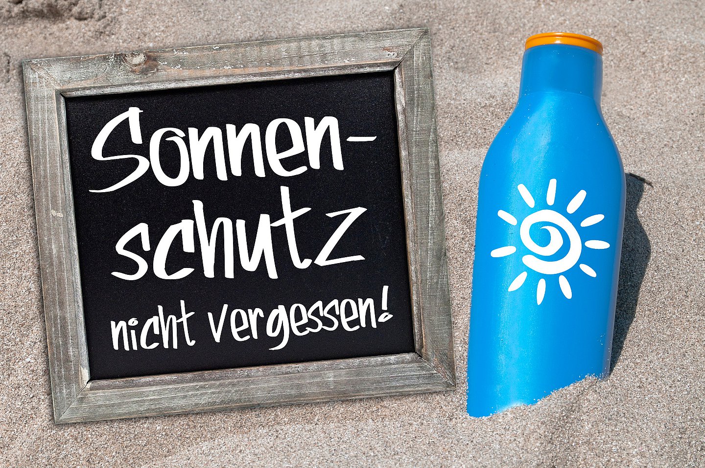 Das Foto zeigt eine Tafel mit dem Hinweis 'Sonnenschutz nicht vergessen' sowie eine Flasche Sonnenmilch am Strand.
