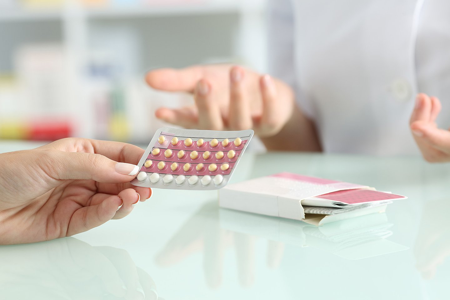 Eine Ärztin im Gespräch mit einer Patientin. Auf einem Tisch liegt eine geöffnete Packung mit einer Antibabypille, von der Patientin sieht man die linke Hand, die einen Blister mit dem Produkt in der Hand hält.