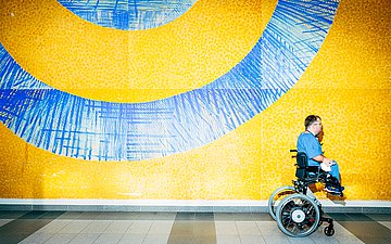 Foto von Charité-Arzt Dr. Leopold Rupp, wie er mit seinem Rollstuhl an einer blau-gelben Wand vorbeifährt