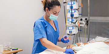 Eine Pflegekraft in blauer Schutzkleidung, mit Handschuhen und OP-Maske, versorgt einen Patienten in einem Krankenhausbett. Sie h&auml;lt einen kleinen Beh&auml;lter mit roter Fl&uuml;ssigkeit in der Hand; im Hintergrund sind medizinische Ger&auml;te und Infusionspumpen zu sehen.
