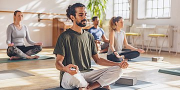 Yoga Das Bild zeigt eine vierköpfige Yoga-Gruppe im Lotussitz. Im Vordergrung ein Mann mit dunklen Haaren, Dutt und Vollbart, im Hintergrund zwei FRauen und ein weiterer Mann.