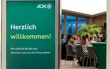 Im linken Bildbereich befindet sich ein gr&uuml;nes Schild mit der Begr&uuml;&szlig;ung "herzlich willkommen!". Im rechten Bildbereich weiter hinten und leicht verschwommen sitzen mehrere Menschen. Sie blicken nach rechts aus dem Bild heraus.