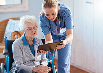Eine &auml;ltere Dame im Rollstuhl; neben ihr steht eine Pflegekraft, die sich zu ihr runterbeugt und ihr etwas in einem Tablet zeigt, das sie in der Hand h&auml;lt.