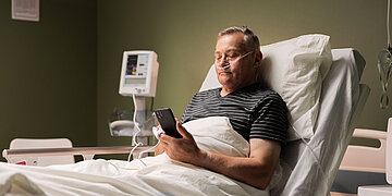 Ein Mann sitzt in einem Krankenhausbett und schaut in sein Handy. In der Nase hat er einen Atemschlauch.