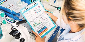 Eine Wissenschaftlerin liest sich Forschungsergebnisse durch. Am Arbeitsplatz sind au&szlig;erdem ein Mikroskop und weitere Unterlagen zu finden.