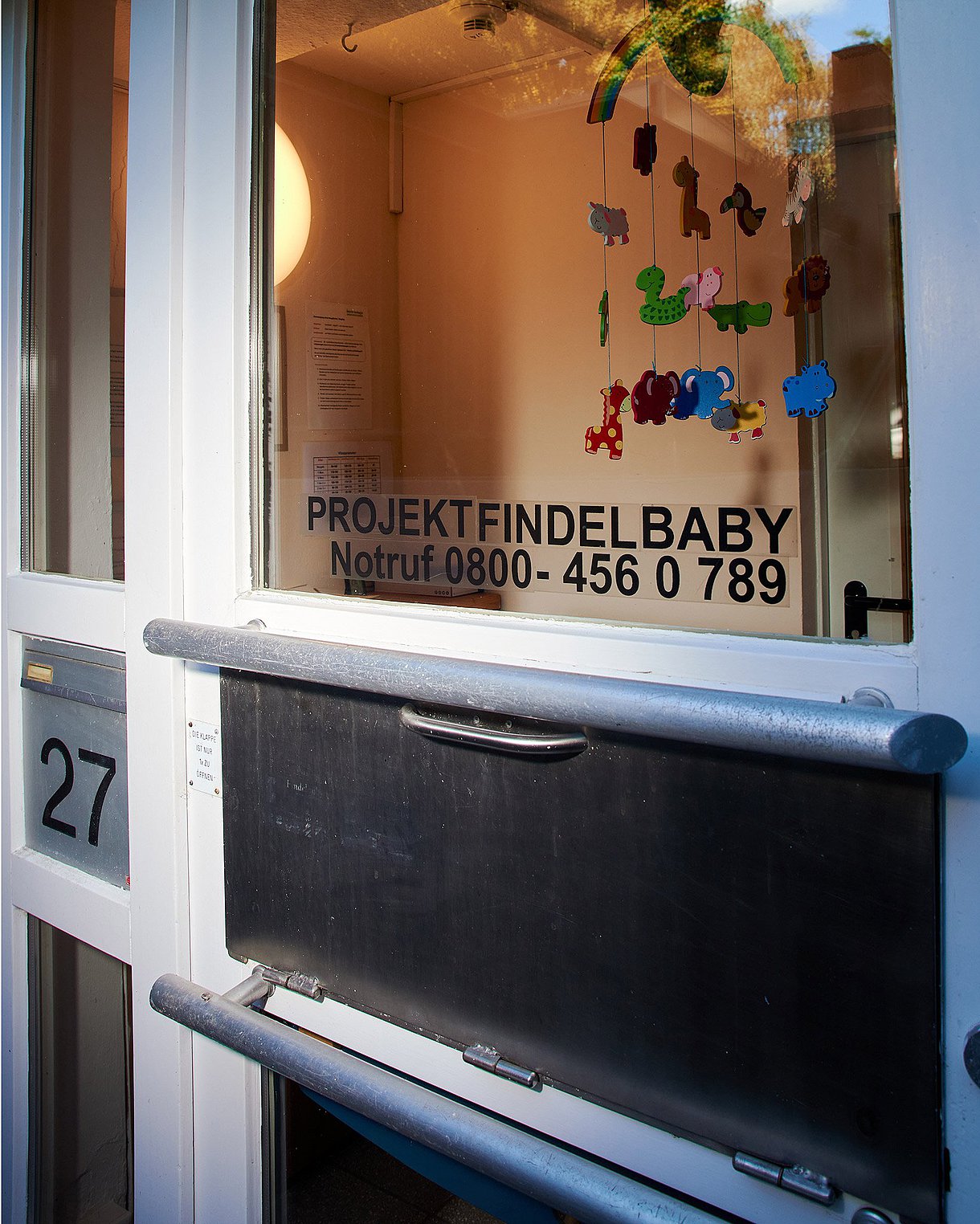 Fotografiert ist die Babyklappe. Eine Vorrichtung mit Griff, in die das Baby beim Projekt Findelbaby reingelegt werden kann. Im Hintergrund erkennt man durch die Scheibe ein buntes Mobilee.