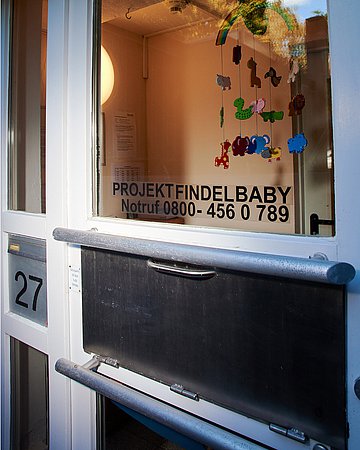 Fotografiert ist die Babyklappe. Eine Vorrichtung mit Griff, in die das Baby beim Projekt Findelbaby reingelegt werden kann. Im Hintergrund erkennt man durch die Scheibe ein buntes Mobilee.