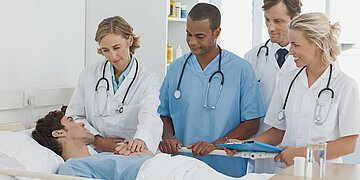 Medizinisch-pflegerisches Personal steht in einem station&auml;ren Umfeld am Bett eines Patienten.