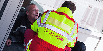 Foto: Eine obdachlose Person lehnt lehnt an einer Wand auf der Stra&szlig;e, davor kniet ein Rettungssanit&auml;ter. 