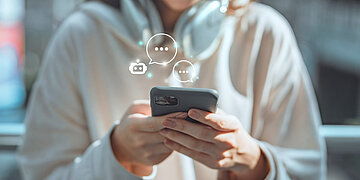 Foto: Eine Frau in hellem Shirt h&auml;lt ein Smartphone in der Hand, dar&uuml;ber angedeutet stehen Sprechblasensymbole.