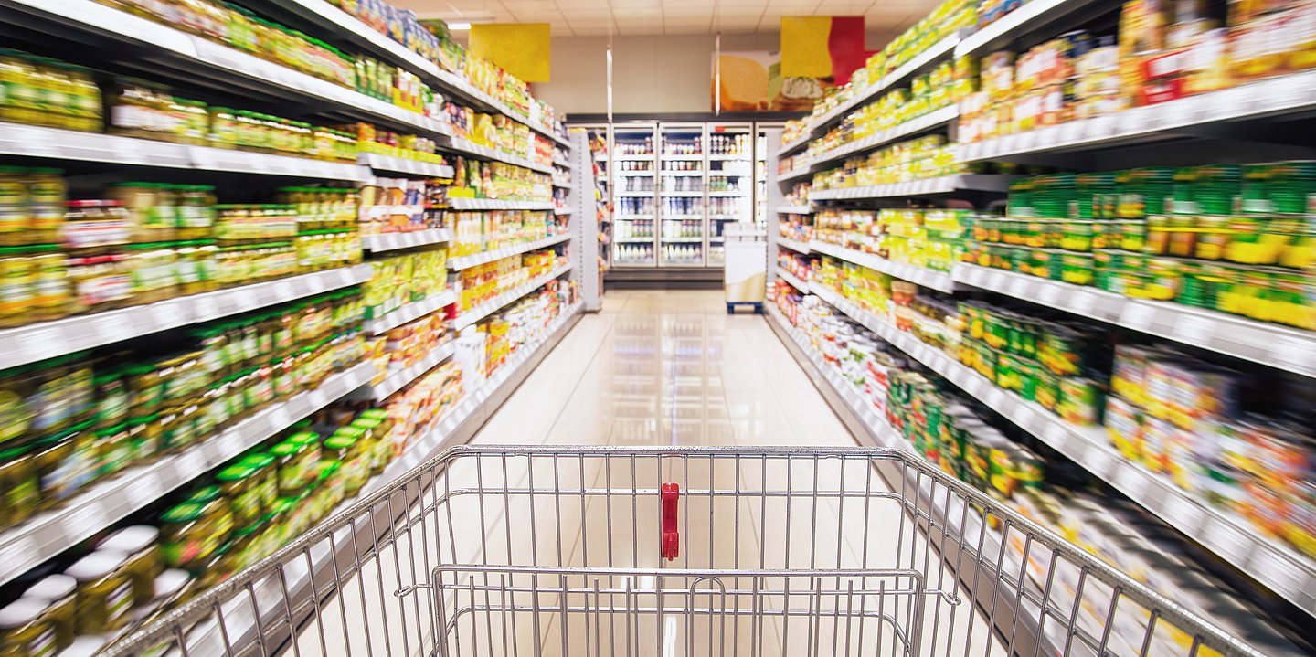 Ein Einkaufswagen rollt zwischen Supermarktregalen mit Fertigprodukten hindurch.