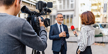 Das Bild zeigt rechts eine Journalistin mit rotbraunen, schulterlangen Haaren und wei&szlig;-schwarz l&auml;ngs gestreifter Bluse. Sie hat die linke Hand in ihrer Hosentasche. In der rechten h&auml;lt sie ein Mikrofon mit rotem Windschutz und interviewt einen Mann mit graumeliertem Haar und Vollbart. Er tr&auml;gt einen graublauen Anzug und ein hellblaues Hemd mit dunkler Krawatte. Im Vordergrund rechts steht ein Kameramann mit grauem Pullover, der die Szenerie mit dem R&uuml;cken zum Betrachter filmt.