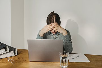 Arbeitsunfähigkeit Eine Frau sitzt an einem Notebook, um zu arbeiten. Ihre Körpersprache zeigt, dass sie sich unwohl fühlt und wahrscheinlich krank wird.