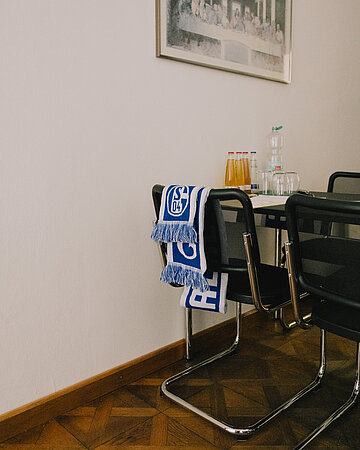 Ein blau-wei&szlig;er Schal des FC Schalke 04 h&auml;ngt &uuml;ber der R&uuml;ckenlehne eines schwarzen Stuhls. Dahinter steht ein Tisch mit Getr&auml;nkeflaschen und Gl&auml;sern in einem hellen Raum mit Parkettboden.