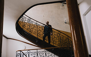 Armin Nassehi steht auf einer geschwungenen Treppe. Das historische Treppenhaus hat ein kunstvoll verziertes, schmiedeeisernes Gel&auml;nder und helle W&auml;nde.
