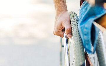 Close-Up zu einem Rollstuhl. Nur das Rad, die Hand am Rad und ein Teil der Sitzfl&auml;che sind zu sehen, auf der eine Person sitzt. Der Rest vom Bild ist Stra&szlig;e.