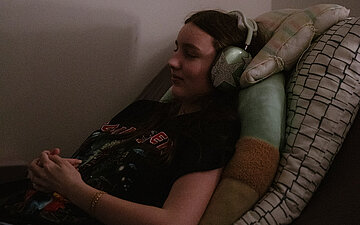Foto: Die 15-j&auml;hrige Elli liegt krankheitsbedingt in ihrem Bett. Noise-Cancelling-Kopfh&ouml;rer schützen sie vor Ger&auml;uschüberlastung.