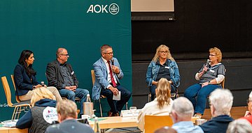 Selbsthilfe in Bad Hersfeld Das Bild zeigt eine Diskussionsrunde aus drei FRauen und einem Mann auf einem Podium plus einem Moderator in der Mitte vor einer dunkelgrünen Wand mit AOK-Logo.. Davor sitzen