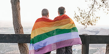Foto: Zwei &auml;ltere M&auml;nner von hinten, gemeinsam in eine Regenbogenflagge geh&uuml;llt, vor einer weiten Landschaft.