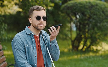 Barrierefreie App Foto eines jungen Mannes, der blind ist und im Freien auf einer Bank sitzt, wo er sein Handy benutzt, um sich etwas vorlesen zu lassen.