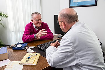 Gespräch Arzt-Patient Das Foto zeigt einen älteren Mann mit weinrotem Pullover, darunter ein farblich passendes gestreiftes Hemd, im Gespräch mit einem Arzt an dessen Schreibtisch. Der Arzthat dem Betrachter den Rücken leicht zugewandt, trägt eine ovale Nickelbrille, trägft einen weißen Kittel und lange Kotletten. Sewine Glatze ist von graumelierten Kurzhaar umkränzt.