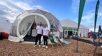 Die AOK auf dem Hessentag 2025 Zwei Männer in blauen Hosen und mit weißem Poloshirt stehen vor einem großen Kugel-Zelt mit großen Fensterflächen. Rechts daneben stehen zwei Strand-Liegestühle mit AOK-Aufschrift und Logo. Rechts dahinter sieht man ein große Veranstaltungszelt