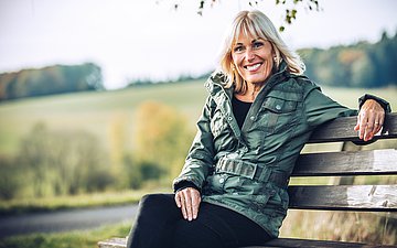 Bettina Müller Das Bild zeigt eine lächelnde blonde Frau mit schulterlangen Haaren. Sie Sitzt unter einem Baum auf einer Bank, trägt einen schwarzen Rock, schwarze Strumpfhosen, ein schwarzes Top und eine grüne Outdoor-Jacke