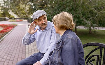 Hören ist wichtig für die Kommunikation. Foto: Zwei Personen im Gespräch auf einer Parkbank, der Mann greift sich an ein Ohr, um besser zu hören.