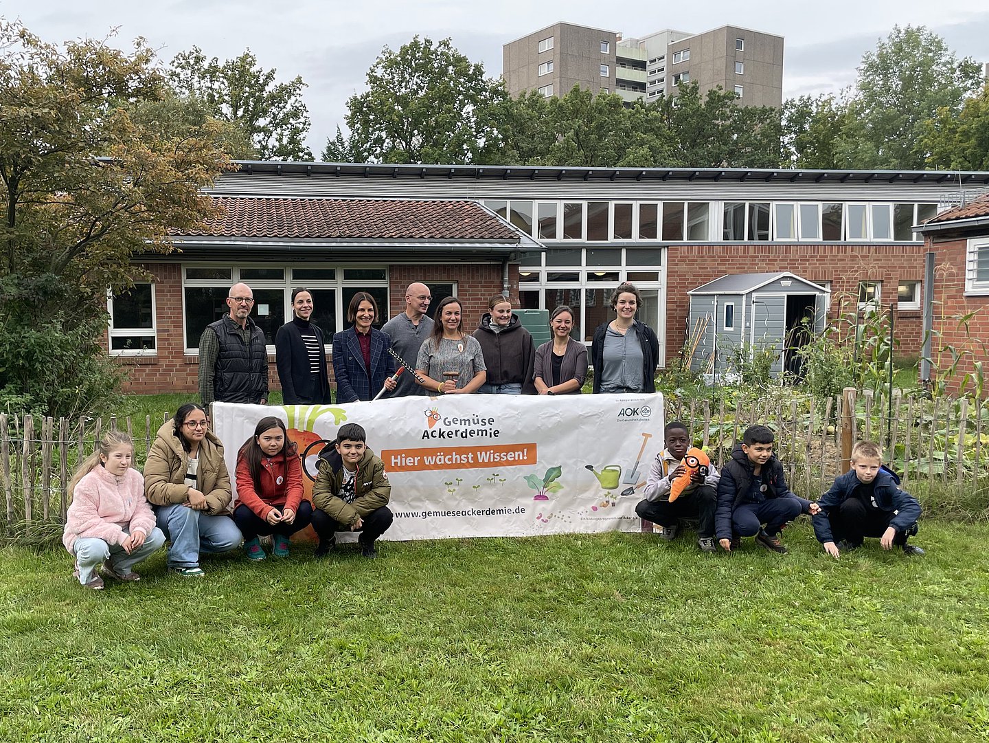 Sandra Kuwatsch, Mitglied des Vorstandes der AOK Niedersachsen und Anouschka Gronau, Geschäftsführerin Gesundheitsmanagement Prävention, AOK Niedersachsen, stehen mit Lehrenden und Mitgliedern der GemüseAckerdemie vor der Grundschule Hägewiesen  hinter einem Banner. Davor sitzen sieben Schülerinnen und Schüler auf dem Rasen.
